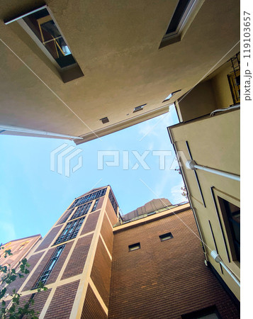 Looking up at the intersection of two modern beige buildings against a bright blue sky. Minimalist urban scene, highlighting the contrast between architecture and open sky 119103657