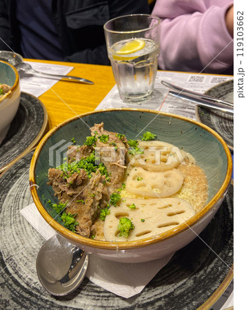 Savory dish with tender meat and lotus root in a rustic bowl, garnished with fresh herbs. intimate dining experience captured, featuring vibrant ingredients and a refreshing glass of water on the side 119103662