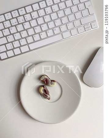 Top view white workspace with keyboard and plate of chocolates. The clean, minimal design emphasizes a blend of work Top view white workspace with keyboard and plate of chocolates. The clean, minimal design emphasizes a blend of work 119103766