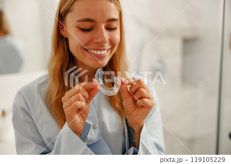 A Smiling Woman Holding a Clear Orthodontic Aligner While Standing in Her Bathroom Setting 119105229