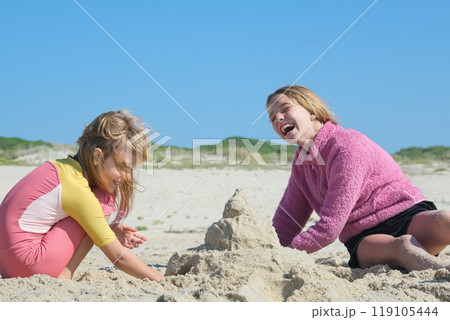 Two girls building a sandcastle on the beach under a clear blue sky. Two happy sisters having fun on the Atlantic ocean deserted beach. Two girls building a sandcastle on the beach under a clear blue sky. Two happy sisters having fun on the Atlantic ocean deserted beach. 119105444