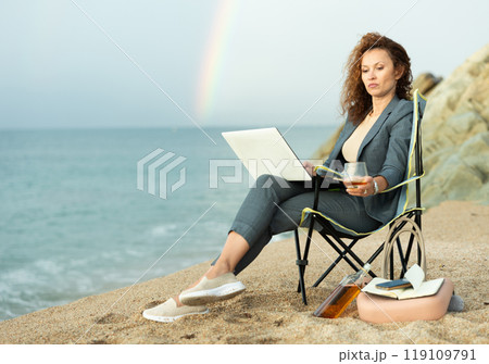 Stylish woman in a suit works on her laptop by the ocean Stylish woman in a suit works on her laptop by the ocean 119109791