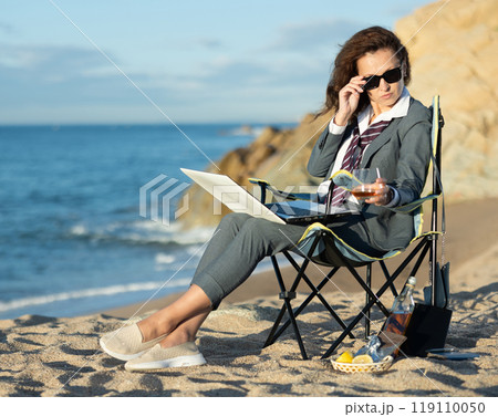 Businesswoman combining work with whiskey by sea on sunny day 119110050