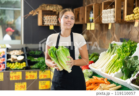 Young female seller in apron displaying assortment of lettuce buds at supermarket 119110057