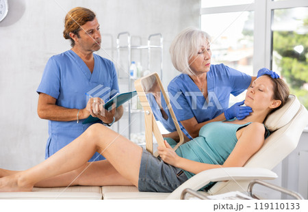 Elderly lady dermatologist examines skin of womans face, male nurse records data in client card Elderly lady dermatologist examines skin of womans face, male nurse records data in client card 119110133