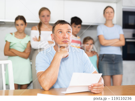 Upset father reads and looks through financial documents. Sad wife with children in background 119110236