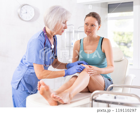 Woman client of clinic looks at specialist and tells to mature woman doctor where knee hurts Woman client of clinic looks at specialist and tells to mature woman doctor where knee hurts 119110297