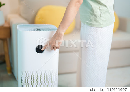 Woman turn on Air Purifier in living room. Purification system for filter and cleaning dust PM2.5 HEPA and virus in home. Allergy, Pure air, health lifestyle, Wellness life and Air Pollution Woman turn on Air Purifier in living room. Purification system for filter and cleaning dust PM2.5 HEPA and virus in home. Allergy, Pure air, health lifestyle, Wellness life and Air Pollution 119111997