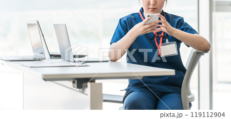 休憩時間にスマホを見るスクラブ姿の医療従事者の女性(顔なし) 休憩時間にスマホを見るスクラブ姿の医療従事者の女性(顔なし) 119112904