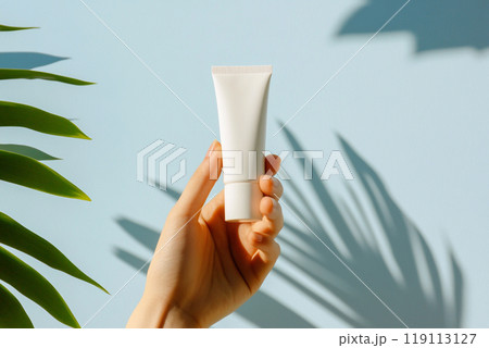 A hand delicately holding a jar of cream, with tropical palm tree leaves framing the sides, set against a serene blue background. skincare and wellness 119113127