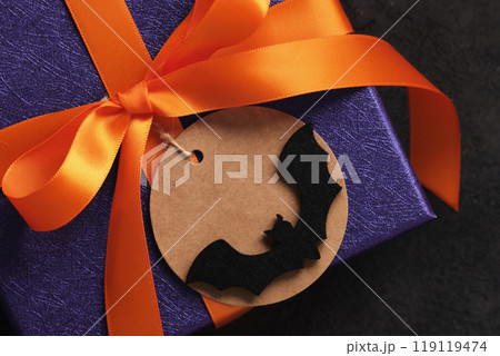 A Halloween gift wrapped in purple paper with orange ribbon and a round tag with a bat silhouette on it 119119474
