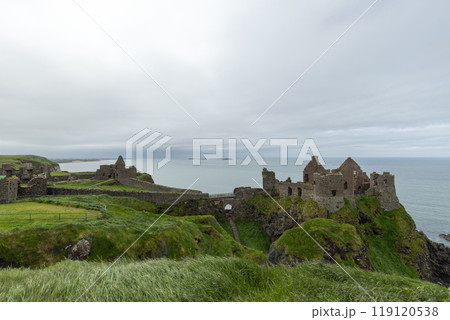 Panoramic view of Dunluce Castle ruins atop a cliff, surrounded by windswept grass and ocean view 119120538