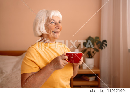Caucasian blonde senior woman sitting on a white bed with a cup in her hand. Female relaxing and drinking cup of hot coffee or tea in the bedroom. 119121994