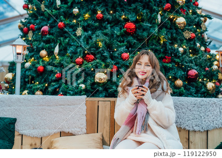 Happy young woman having Hot Drink and relaxing in lounge zone on traditional city street Christmas market. Festive mood at Xmas fair of European city. Winter holidays, Merry Xmas and Happy New Year. Happy young woman having Hot Drink and relaxing in lounge zone on traditional city street Christmas market. Festive mood at Xmas fair of European city. Winter holidays, Merry Xmas and Happy New Year. 119122950
