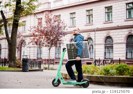 Man rides electric scooter while carrying solar panel. Integration of solar power as sustainable energy source for charging electric scooters, promoting eco-friendly urban transportation. Man rides electric scooter while carrying solar panel. Integration of solar power as sustainable energy source for charging electric scooters, promoting eco-friendly urban transportation. 119125070