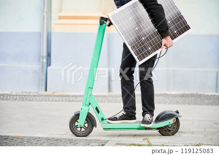 Cropped view of man rides electric scooter while carrying solar panel. Integration of solar power as sustainable energy source for charging electric scooters, promoting eco-friendly transportation. 119125083