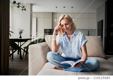 Woman sitting comfortably on couch, deeply engaged in reading book. Cozy living space with modern aesthetic on background. Person enjoying quiet moment of solitude, immersed in reading. Woman sitting comfortably on couch, deeply engaged in reading book. Cozy living space with modern aesthetic on background. Person enjoying quiet moment of solitude, immersed in reading. 119125085