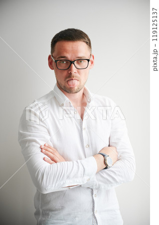 Portrait of man sticking his tongue out in a playful manner. Handsome young male in casual clothes keeping arms crossed while standing on white background. 119125137