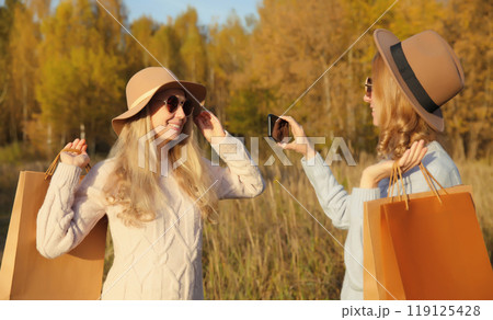 Happy women friends taking selfie with phone, shopping bags in autumn, girlfriends smiling in park Happy women friends taking selfie with phone, shopping bags in autumn, girlfriends smiling in park 119125428