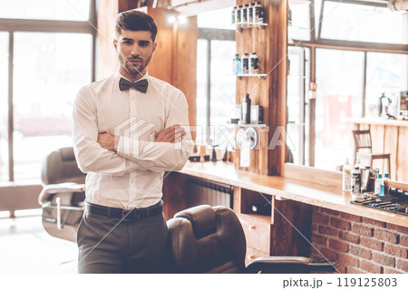 Loving his job. Young handsome man looking at camera and keeping arms crossed while standing at barbershop Loving his job. Young handsome man looking at camera and keeping arms crossed while standing at barbershop 119125803
