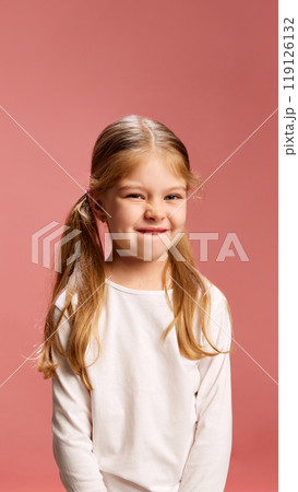 Smiling softly, girl bites her lip in shy but playful gesture. Sweet and innocent moment against pastel pink background. 119126132