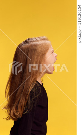 Side profile of girl screaming loudly, her mouth wide open,against yellow studio background, as if in heated tantrum or letting out all her frustrations 119126154