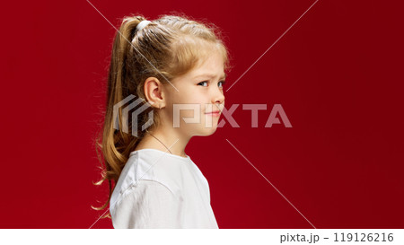 Little girl looks deeply thoughtful, her hand resting under her chin in classic thinking pose, as though pondering something important against red background. 119126216