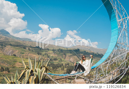 A girl sits on a blue viewpoint in a field with mountains in the background, traveling through the peruvian andes. Canta. Lima, Peru. 119126834