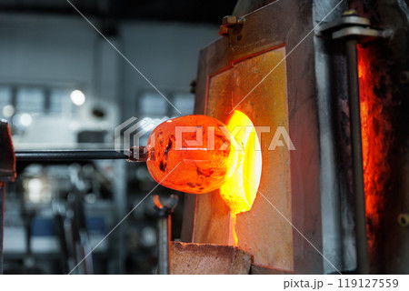 Close-up glassblowing process tube tool with molten glass shaped inside refractory furnace in industrial craft workshop. Handmade blowing glassware design production manufacture Europe 119127559