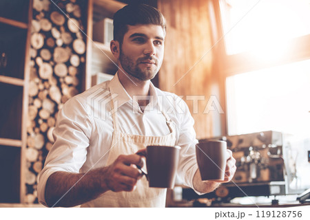 Here you go! Young handsome man in apron holding two coffee cups and looking away while standing at cafe 119128756