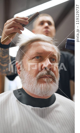 Barber shaping man hair with round brush and hairdryer, emphasizing detail and technique. Elderly man making hairstyle in barber shop 119129737