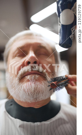 Close-up of elderly man with eyes closed, having beard grooming. Brush and hairdryer used to shape his beard. Barbershop attendance 119129739