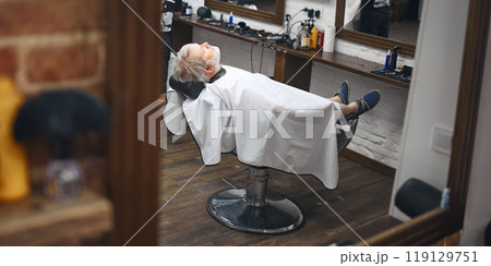 Client reclining in chair, fully relaxed, showcasing the comfort. Elderly man visiting barbershop for hairstyle 119129751
