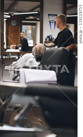Barber leaning over reclined client, carefully trimming his beard. Client appears comfortable and relaxed in reclining chair 119129753