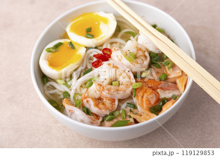 Asian dish with broth, noodles, tiger shrimp, fish, egg and green onions. Traditional Asian soup in a white deep plate. Selective focus, close-up. Asian dish with broth, noodles, tiger shrimp, fish, egg and green onions. Traditional Asian soup in a white deep plate. Selective focus, close-up. 119129853