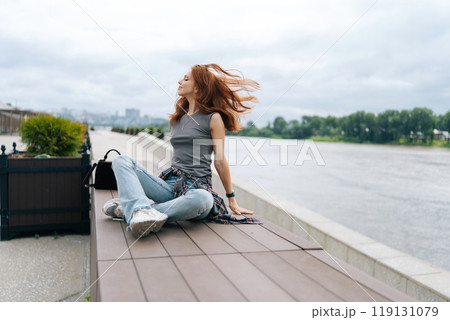 Carefree young woman sitting on bench with hair blowing in wind, enjoying fresh air and view of river on cloudy summer day. Pretty smiling redhead female appears at ease, taking respite alone. 119131079