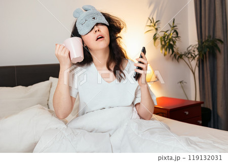 Portrait of funny sleeping young woman wearing sleep mask call with cup and drink with phone while sitting in bed. Concept of being late for work and morning awakening Portrait of funny sleeping young woman wearing sleep mask call with cup and drink with phone while sitting in bed. Concept of being late for work and morning awakening 119132031
