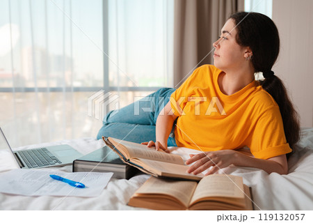 Portrait of young caucasian woman is lying on her bed and holding a book. Laptop and documents are lying next to each other. The concept of online courses and online education 119132057