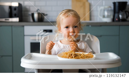 Delightful baby sits in a high chair, determined to eat homemade pasta bolognese with a fork in a cozy kitchen 119132147