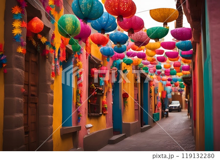 Colorful metallic paper pinatas suspended on public street between houses and buildings of colonial town against blue sky in background, Mexican tradition 119132280