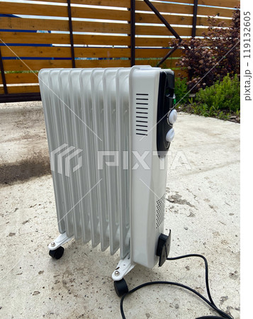 White oil-filled radiator stands outside on a concrete surface, prepared to provide warmth on a chilly day. Portable design makes it a practical heating solution for various environments 119132605