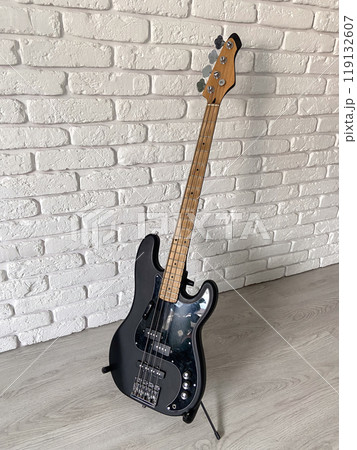 Sleek black electric bass guitar stands poised against a textured white brick wall, capturing the essence of modern music and style. The contrast between the dark instrument and the light surroundings 119132607