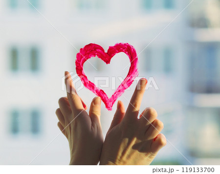 Fingers draw red heart sign on the window glass in sunlight. The concept of Valentine's Day, romantic relationships, Gift Red heart emotional. Closing feelings, lonelyness. Revealing feelings, trust. 119133700