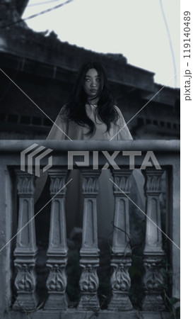 Woman ghost standing behind balustrade. Woman had long hair. The sky is dark and cloudy, and there is a abandoned building in the background. Scene is scary and creepy 119140489