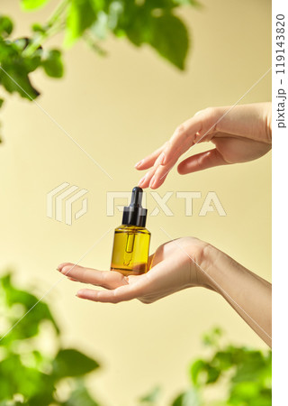 A mockup of a yellow glass bottle with a dropper that holds an essential is carefully held by a model hand in the surrounding greenery and the whitish background of the picture. A mockup of a yellow glass bottle with a dropper that holds an essential is carefully held by a model hand in the surrounding greenery and the whitish background of the picture. 119143820