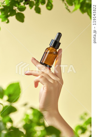 The image features a hand delicately holding a brown glass bottle with a dropper mockup, containing an essential inside. The yellow background and surrounding green foliage. 119143822
