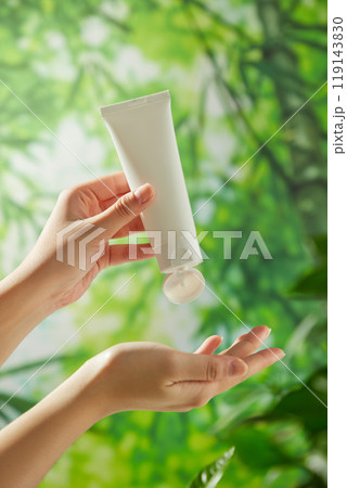 A template image for an ad features a lush bamboo forest backdrop with a model hand holding a white tube without a label and squeezing a cream sample into the other hand to show off a creamy texture. 119143830