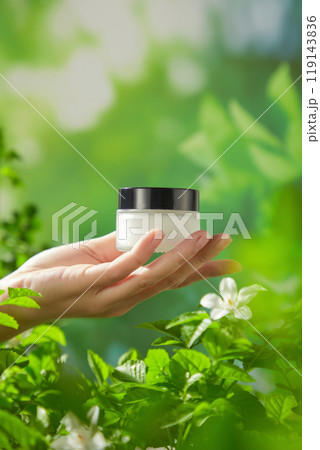 A blurred green foliage background features a model holding a small glass jar with the black cap in the center of the image, perfect for promoting natural and high-quality skincare products. 119143836