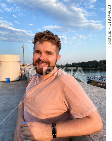 Bearded man gazes thoughtfully at the sunset, leaning against a concrete railing on a scenic promenade. Soft glow of the setting sun creates a serene and reflective atmosphere Bearded man gazes thoughtfully at the sunset, leaning against a concrete railing on a scenic promenade. Soft glow of the setting sun creates a serene and reflective atmosphere 119144623