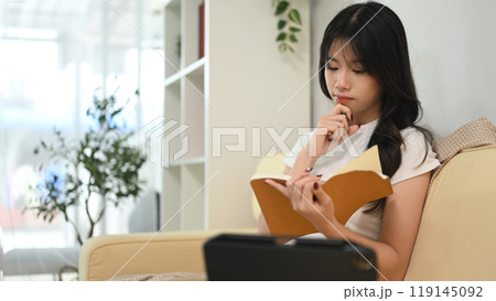 Peaceful young woman sitting on beige sofa and reading book Peaceful young woman sitting on beige sofa and reading book 119145092